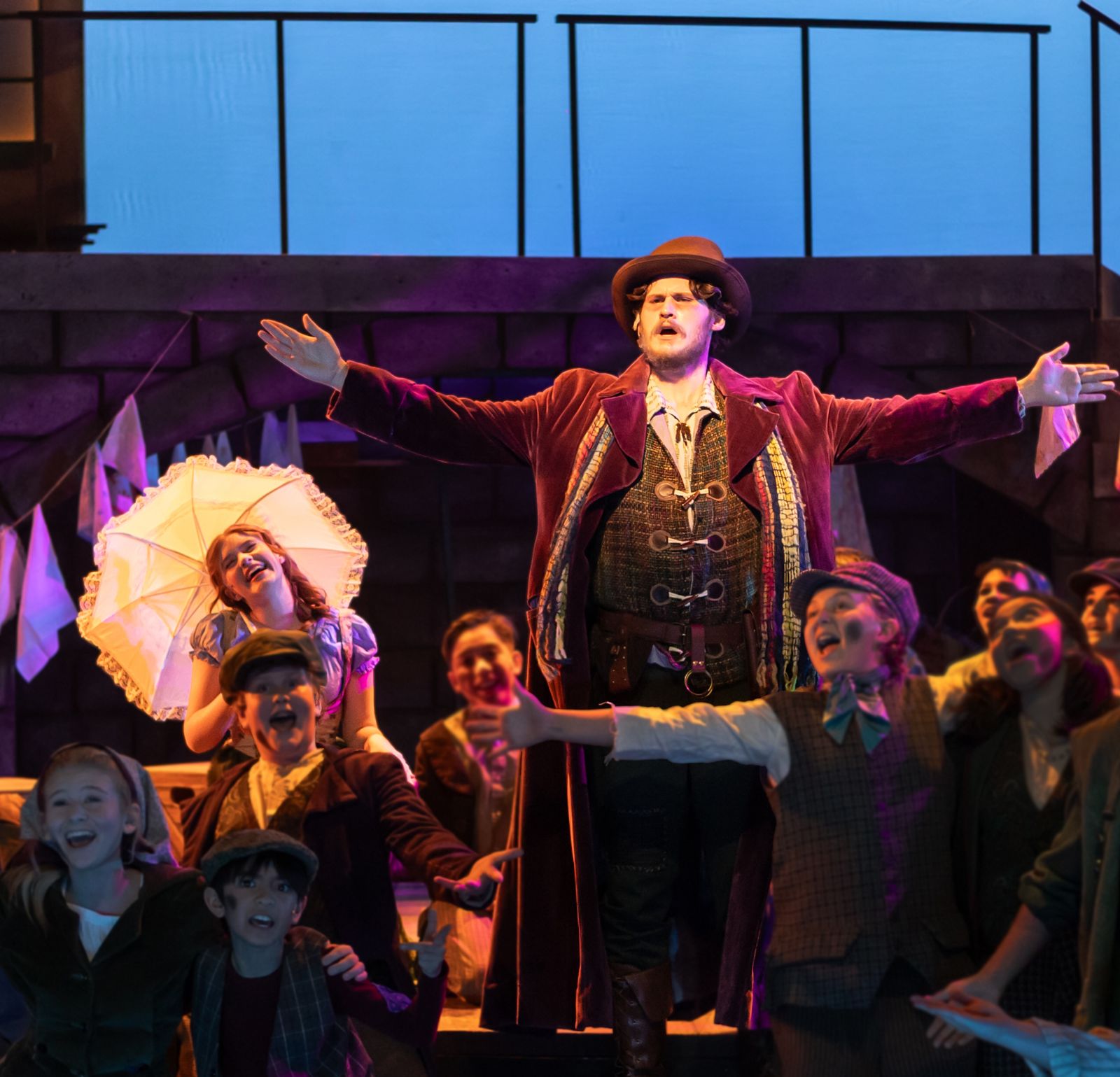 Parker Pitt as Bet holding a white parasol behind Fagin, arms spread, with the company in Oliver! at TheaterWorks